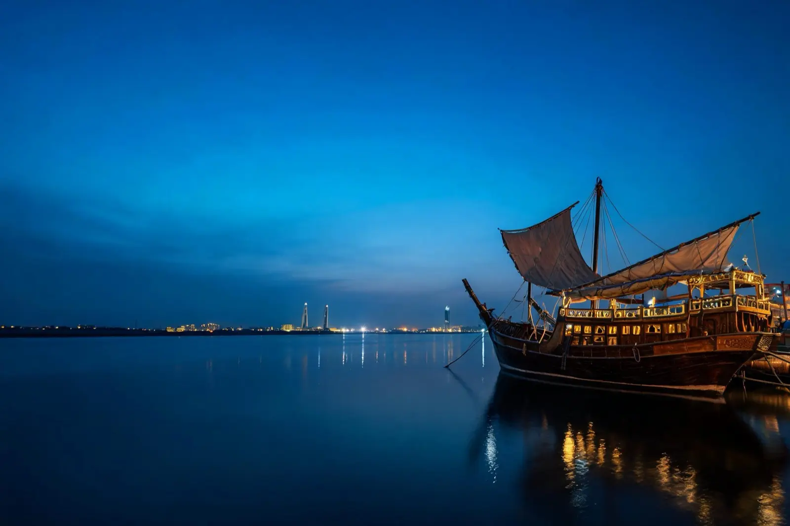Hölzerne Dhau mit Segeln in einem ruhigen Hafen, am Horizont die Lichter einer Küstenstadt (KI-Illustration)